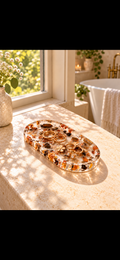Oval clear-look decorative jewellery tray with warm earth-toned details on a sunlit vanity; jewellery shown for styling.